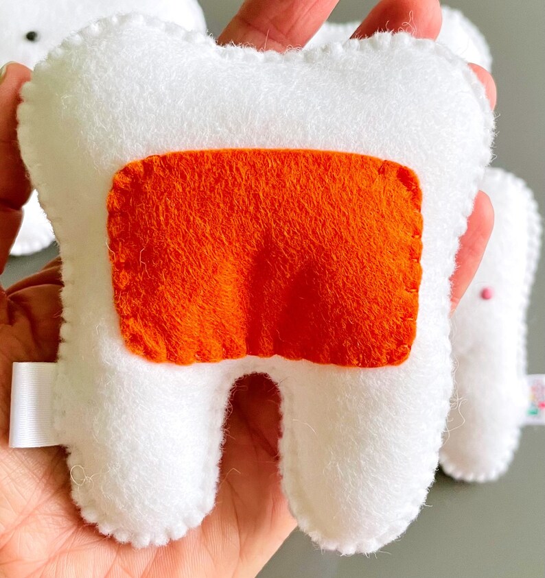 May include: White felt tooth with a bright orange felt patch on the front. The tooth is a plush toy.