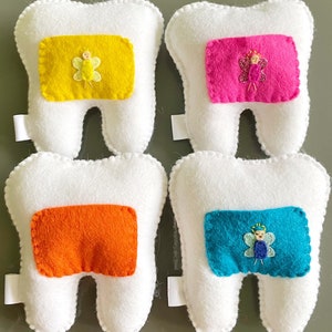 May include: Four white felt tooth pillows with different colored felt patches. Each patch features a small embroidered fairy with wings. The colors of the patches are yellow, pink, orange, and blue.