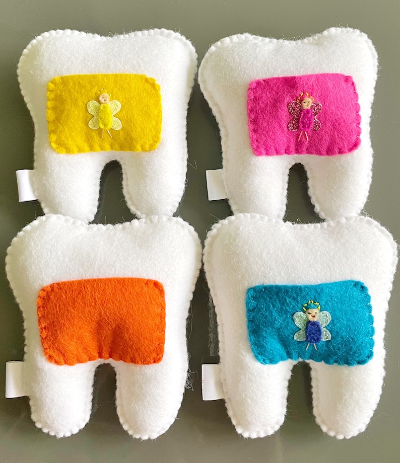 May include: Four white felt tooth pillows with different colored felt patches. Each patch features a small embroidered fairy with wings. The colors of the patches are yellow, pink, orange, and blue.