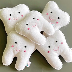 May include: Five white plush teeth with embroidered smiling faces. Each tooth has black eyes and pink cheeks. The teeth are arranged in a pile.
