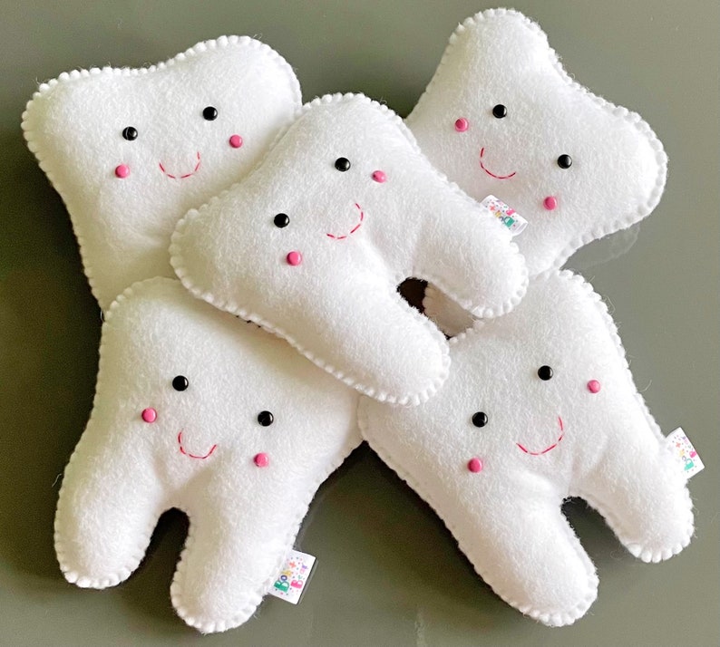 May include: Five white plush teeth with embroidered smiling faces. Each tooth has black eyes and pink cheeks. The teeth are arranged in a pile.