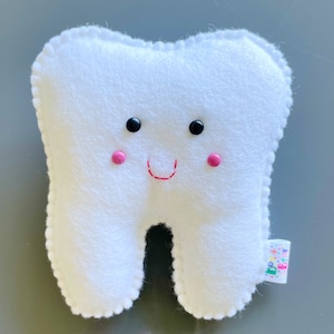 May include: White plush tooth with a smiling face. The tooth has black eyes, pink cheeks, and a red stitched smile.