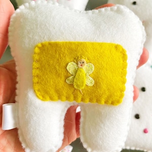 May include: A white felt tooth with a yellow felt pocket on the front. The pocket has a small embroidered fairy with a yellow dress and wings.