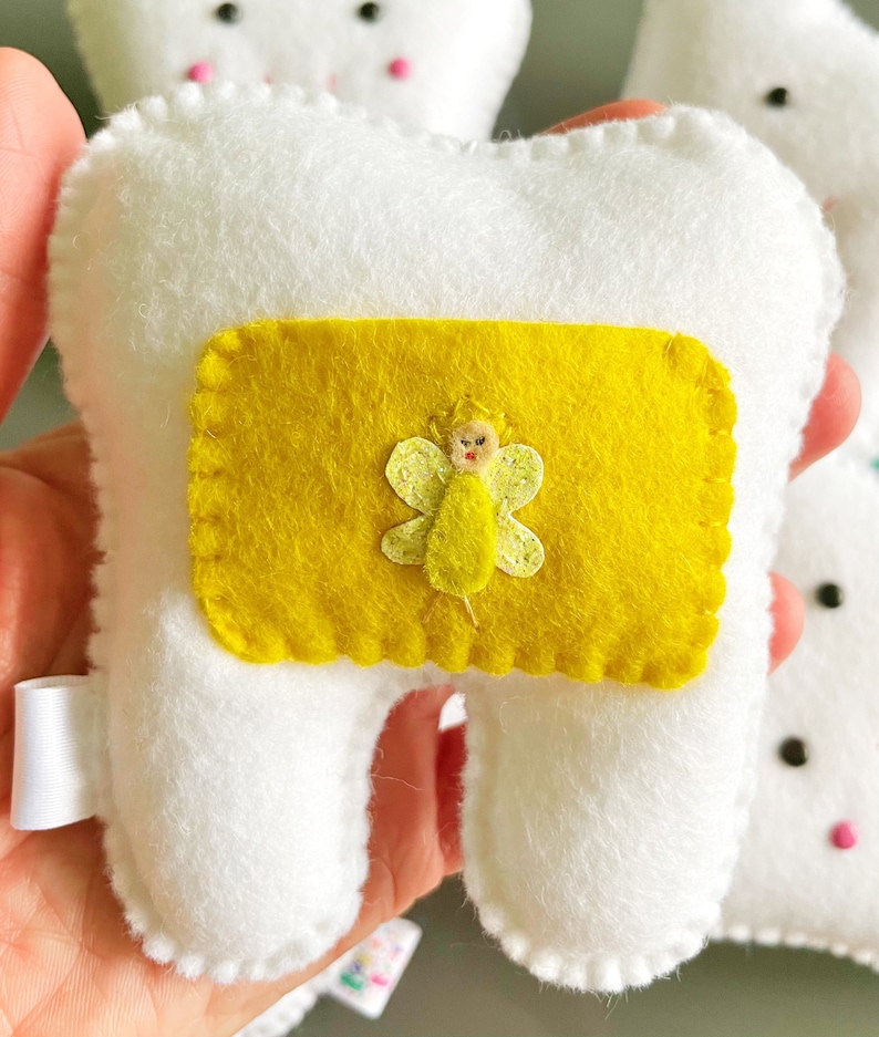 May include: A white felt tooth with a yellow felt pocket on the front. The pocket has a small embroidered fairy with a yellow dress and wings.