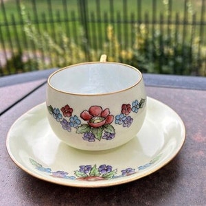 May include: Cream-colored teacup and saucer set. The cup and saucer are decorated with a floral pattern featuring red, blue, and purple flowers. The rims are trimmed with gold. The set is placed on a dark surface.