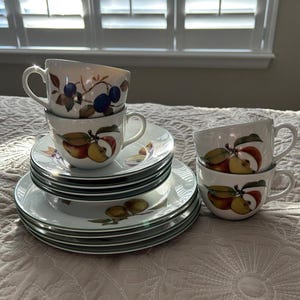 May include: A set of four white porcelain teacups and saucers with a floral pattern featuring apples and plums. The cups have a delicate handle and the saucers have a scalloped edge.