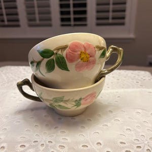 May include: Two vintage teacups with a floral pattern of pink roses and green leaves. The cups are stacked on top of each other and have a light brown colour.