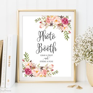 May include: A framed sign with the words "Photo Booth" in elegant script, adorned with watercolor floral arrangements in pink and burgundy. The sign also includes the text "Grab a Prop and Strike a Pose."