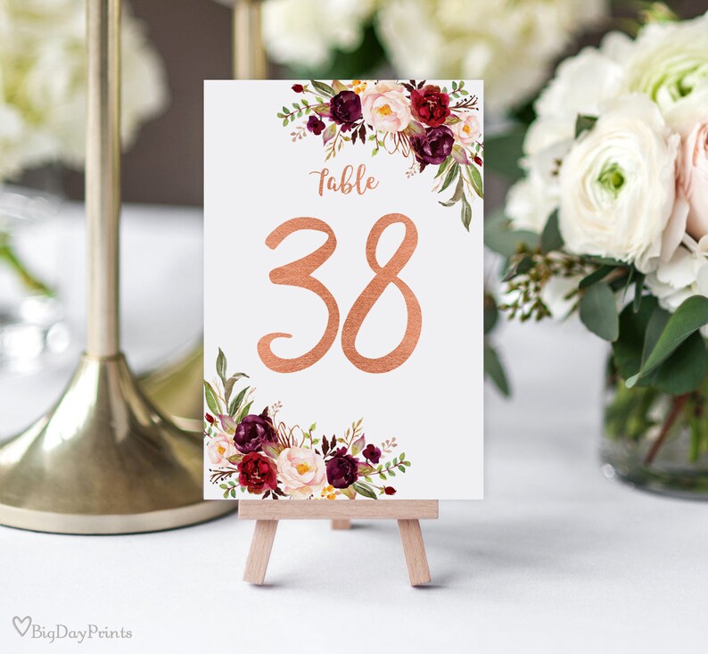 May include: A white table number card with the number 38 in copper script. The card is decorated with floral arrangements in burgundy, cream, and green. The card is displayed on a small wooden easel.