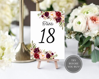 May include: A white table number card with the number 18 in large black font, accented with floral arrangements in burgundy, pink, and green. The card is displayed on a small wooden easel, with a "Try Before You Buy!" sticker.