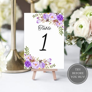 May include: Purple and white floral wedding table number card with the text "Table 1" on a small wooden easel.