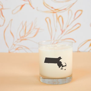 May include: A clear glass candle holder filled with white wax. A black silhouette of the state of Massachusetts is printed on the front of the candle. The background features a white wall with orange floral designs.