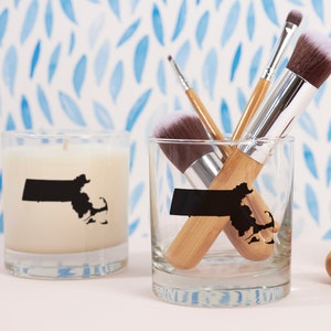 May include: A candle and a glass with a black Massachusetts state outline. The glass contains makeup brushes with wooden handles and white bristles. The candle is white and the glass is clear.
