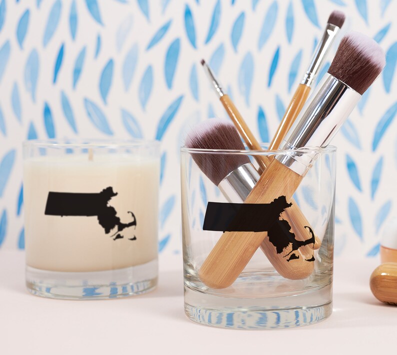 May include: A candle and a glass with a black Massachusetts state outline. The glass contains makeup brushes with wooden handles and white bristles. The candle is white and the glass is clear.