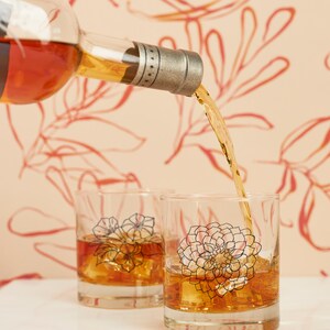 May include: A bottle pours amber liquid into two clear, lowball glasses filled with ice. Each glass features a floral design in black and pink. The background is a light peach color with a red floral pattern.