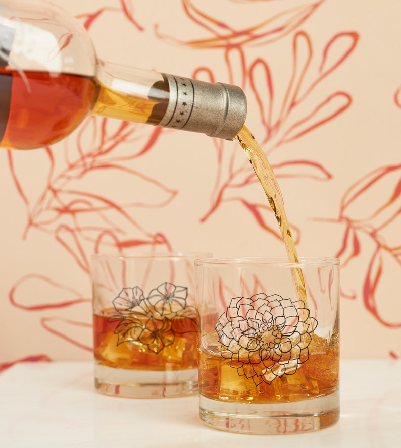 May include: A bottle pours amber liquid into two clear, lowball glasses filled with ice. Each glass features a floral design in black and pink. The background is a light peach color with a red floral pattern.