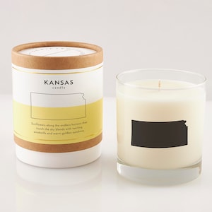 K&ouml;nnte beinhalten: Ein wei&szlig;es Kerzengef&auml;&szlig; mit einer schwarzen Silhouette des Bundesstaates Kansas. Das Gef&auml;&szlig; hat ein gelbes Etikett mit dem Text "KANSAS candle" und dem Satz "Sunflowers along the endless horizon that touch the sky blends with twirling windmills and warm golden sunshine."