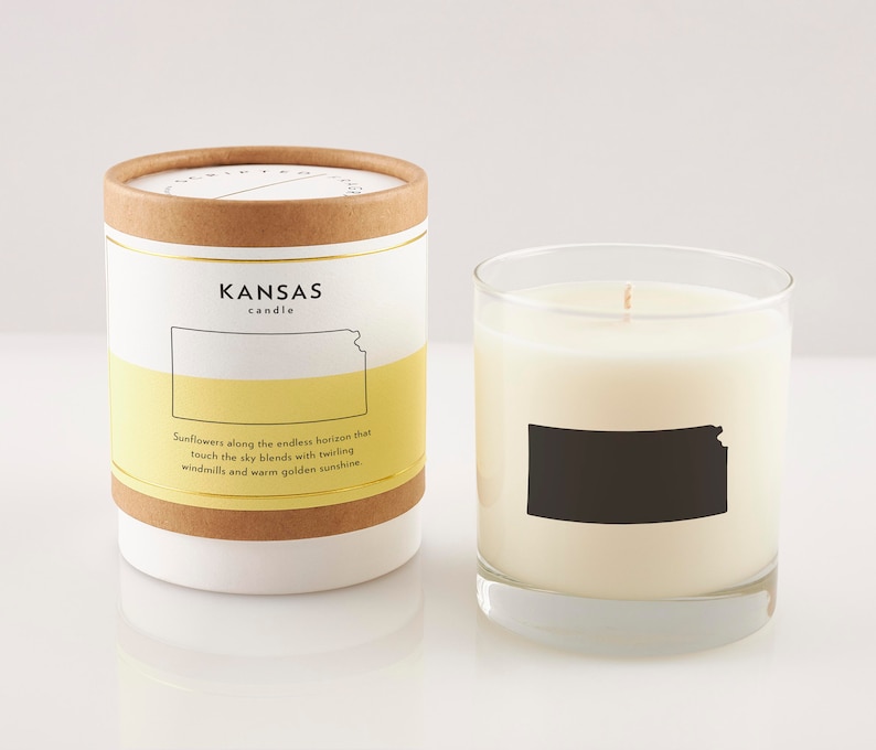 K&ouml;nnte beinhalten: Ein wei&szlig;es Kerzengef&auml;&szlig; mit einer schwarzen Silhouette des Bundesstaates Kansas. Das Gef&auml;&szlig; hat ein gelbes Etikett mit dem Text "KANSAS candle" und dem Satz "Sunflowers along the endless horizon that touch the sky blends with twirling windmills and warm golden sunshine."