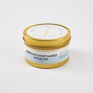 May include: A gold-colored metal candle tin with a white label that reads "Scripted Fragrance" and "German Shorthaired Pointer candle." The candle has a light blue band with text describing the scent. The candle is on a white background.