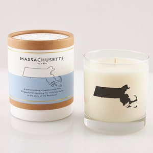 May include: A white candle in a clear glass with a black silhouette of Massachusetts. The candle is next to a cylindrical container with the text "MASSACHUSETTS candle" and a map outline. The container has a blue band with text.