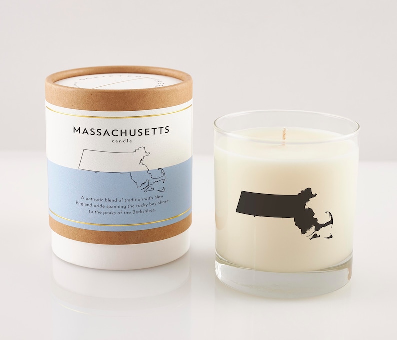 May include: A white candle in a clear glass with a black silhouette of Massachusetts. The candle is next to a cylindrical container with the text "MASSACHUSETTS candle" and a map outline. The container has a blue band with text.