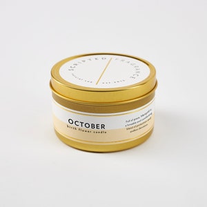May include: A gold-colored candle tin with the text "SCRIPTED FRAGRANCE" and "OCTOBER birth flower candle" printed on the label. The candle has a white and beige label with additional text. The candle is on a white background.
