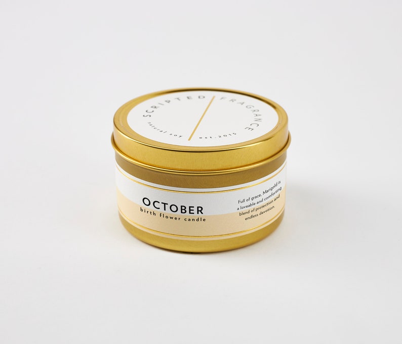 May include: A gold-colored candle tin with the text "SCRIPTED FRAGRANCE" and "OCTOBER birth flower candle" printed on the label. The candle has a white and beige label with additional text. The candle is on a white background.