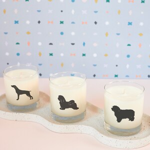 May include: Three glass candles with dog silhouettes on a speckled white tray. Each candle has a different dog breed silhouette in black. The background is a light blue with colorful geometric shapes. The candles are unscented.