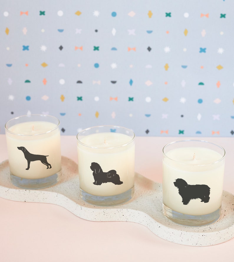 May include: Three glass candles with dog silhouettes on a speckled white tray. Each candle has a different dog breed silhouette in black. The background is a light blue with colorful geometric shapes. The candles are unscented.