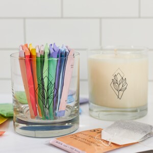 May include: A clear glass tumbler filled with colorful tea packets, next to a lit candle in a clear glass. Both items feature a black geometric leaf design. Tea bags are scattered around the candle and tumbler.