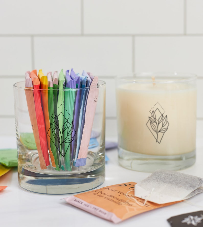 May include: A clear glass tumbler filled with colorful tea packets, next to a lit candle in a clear glass. Both items feature a black geometric leaf design. Tea bags are scattered around the candle and tumbler.
