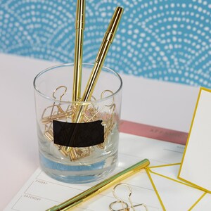 K&ouml;nnte beinhalten: Ein Glasgef&auml;&szlig; gef&uuml;llt mit goldenen B&uuml;roklammern und zwei goldenen Stiften. Das Gef&auml;&szlig; hat ein schwarzes Etikett mit der Aufschrift "To Do". Das Gef&auml;&szlig; steht auf einem wei&szlig;en Schreibtisch mit einem rosa und gelben Planer und einer wei&szlig;en Karte.