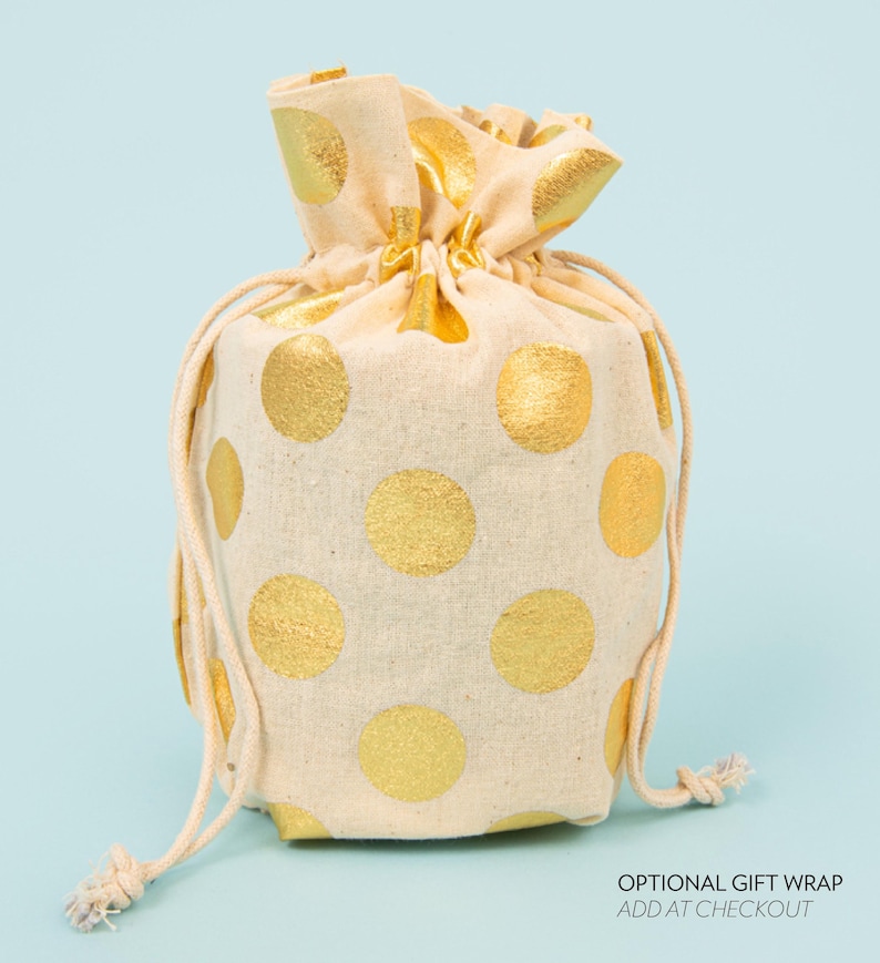 May include: A beige drawstring gift bag with large gold polka dots. The bag is tied at the top with a beige drawstring. The text "OPTIONAL GIFT WRAP ADD AT CHECKOUT" is visible at the bottom right.