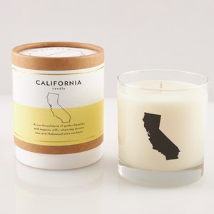 May include: A California candle in a clear glass and a matching cylindrical container. The candle and container feature a black silhouette of California. Text on the container reads "CALIFORNIA candle" and "A sun-kissed blend of golden beaches and majestic cliffs, where big dreams soar and Hollywood stars are born."