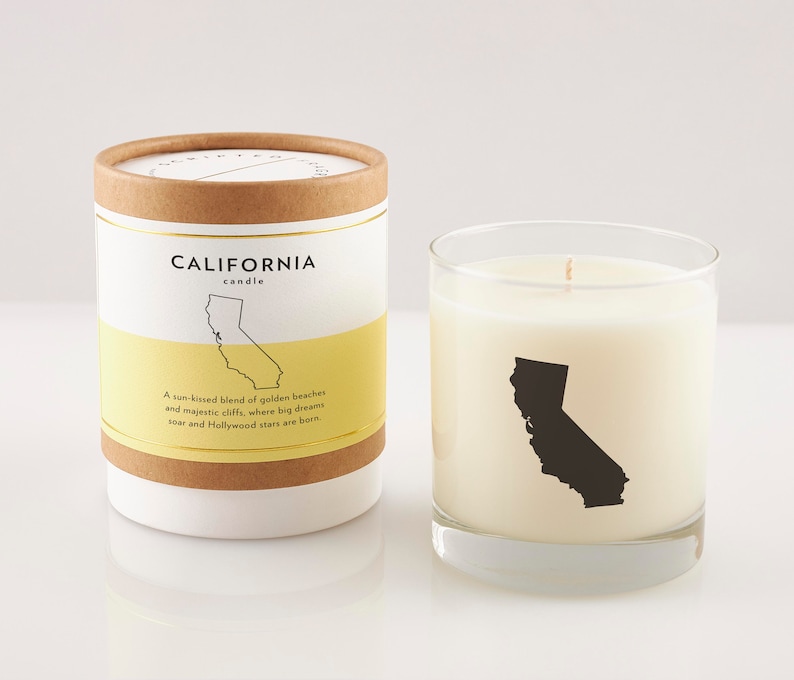 May include: A California candle in a clear glass and a matching cylindrical container. The candle and container feature a black silhouette of California. Text on the container reads "CALIFORNIA candle" and "A sun-kissed blend of golden beaches and majestic cliffs, where big dreams soar and Hollywood stars are born."