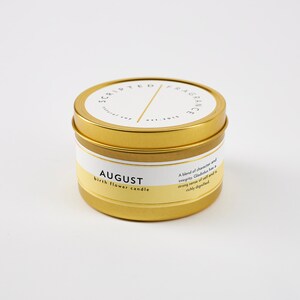 Op de afbeelding: Een goudkleurige kaarsenblik met een wit en geel etiket. Op het etiket staat "AUGUST birth flower candle" en "SCRIPTED FRAGRANCE". De kaars is een natuurlijke sojakaars.
