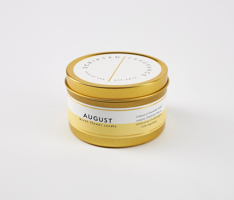 Op de afbeelding: Een goudkleurige kaarsenblik met een wit en geel etiket. Op het etiket staat "AUGUST birth flower candle" en "SCRIPTED FRAGRANCE". De kaars is een natuurlijke sojakaars.