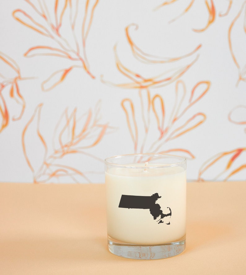 May include: A clear glass candle holder filled with white wax. A black silhouette of the state of Massachusetts is printed on the front of the candle. The background features a white wall with orange floral designs.