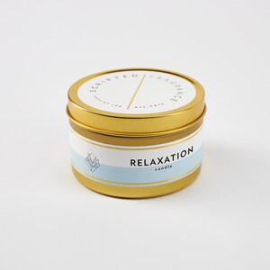 May include: A gold-colored candle tin with a white and light blue label. The label reads "RELAXATION candle" and "SCRIPTED FRAGRANCE est. 2015". The candle tin has a gold lid and a gold band around the middle.