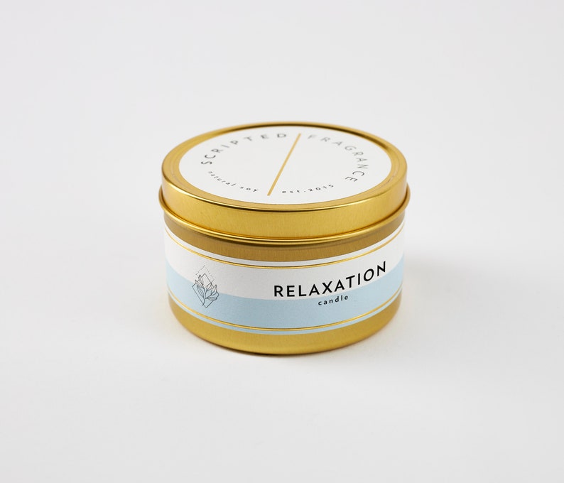 May include: A gold-colored candle tin with a white and light blue label. The label reads "RELAXATION candle" and "SCRIPTED FRAGRANCE est. 2015". The candle tin has a gold lid and a gold band around the middle.