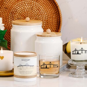 May include: A collection of white candles, jars, and a glass with a bridge design. One candle has a gold heart, another has a bridge graphic, and the third has the text "Newport". The items are arranged on a white surface with a woven basket in the background.