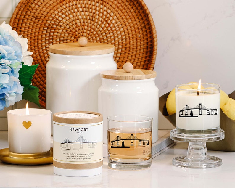 May include: A collection of white candles, jars, and a glass with a bridge design. One candle has a gold heart, another has a bridge graphic, and the third has the text "Newport". The items are arranged on a white surface with a woven basket in the background.