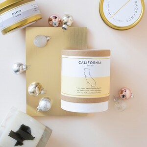 May include: An overhead shot of several scented candles with gold lids and labels, including one labeled "Ohio" and another "California." Small, shiny ornaments in silver and rose gold are scattered around the candles.