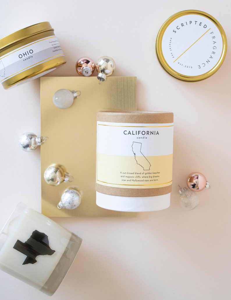 May include: An overhead shot of several scented candles with gold lids and labels, including one labeled "Ohio" and another "California." Small, shiny ornaments in silver and rose gold are scattered around the candles.