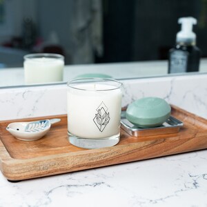May include: A wooden tray holds a clear glass candle with a black geometric design, a small ceramic bird dish, and a green soap on a metal dish. The candle is white, and the soap is round.