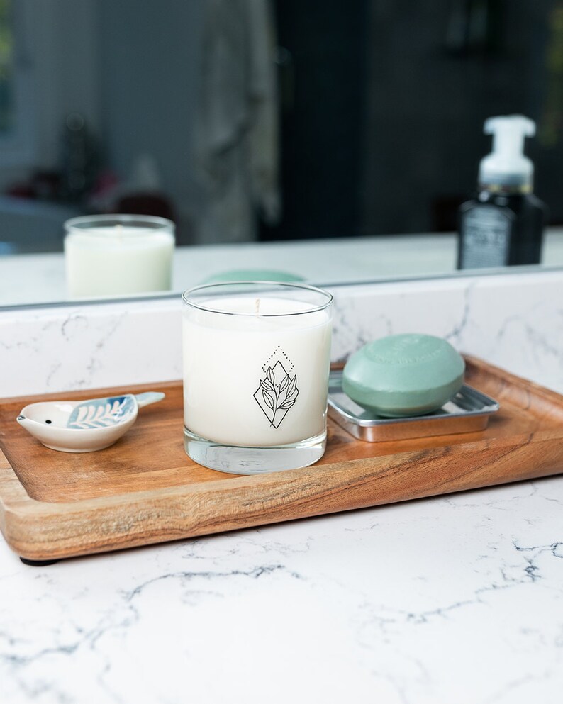 May include: A wooden tray holds a clear glass candle with a black geometric design, a small ceramic bird dish, and a green soap on a metal dish. The candle is white, and the soap is round.