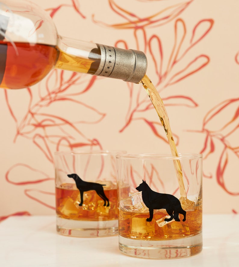 May include: Two clear whiskey glasses with black dog silhouettes, filled with ice and amber liquid, are being poured into from a bottle. The background features a peach-colored wall with a red floral pattern.