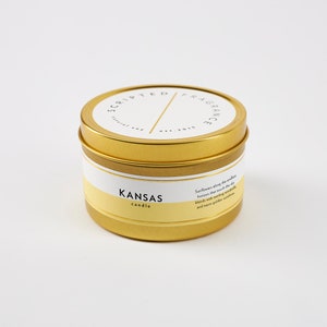 K&ouml;nnte beinhalten: Goldene Kerzen-Dose mit einem wei&szlig;en Etikett, auf dem "Scripted Fragrance" und "Kansas candle" steht. Das Etikett beschreibt den Duft auch als "Sonnenblumen entlang des endlosen Horizonts, die den Himmel ber&uuml;hren, vermischen sich mit funkelnden Gl&uuml;hw&uuml;rmchen und warmer goldener Sonne."