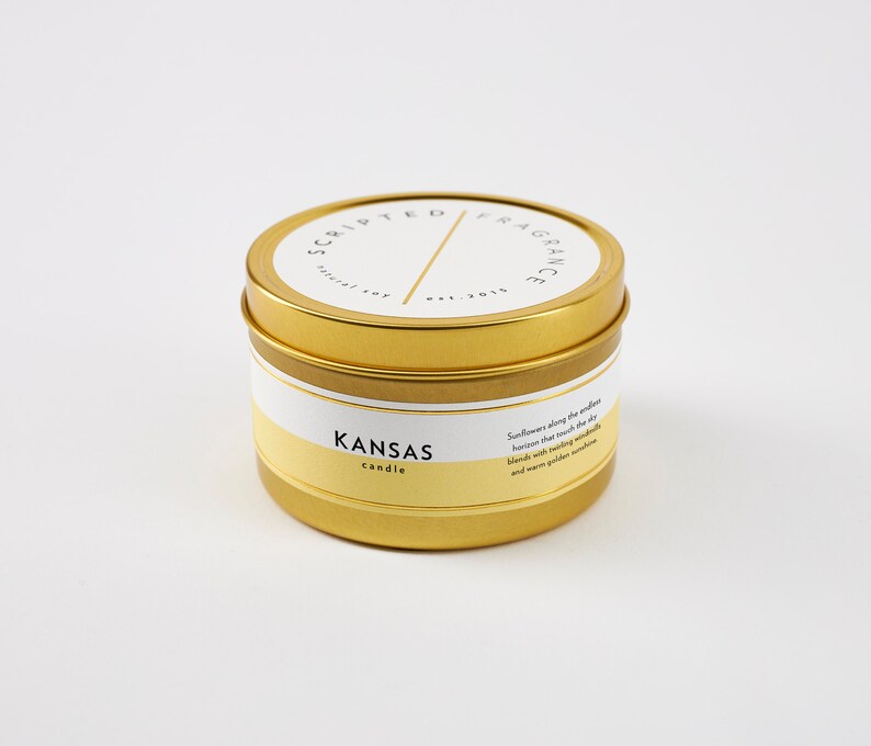 K&ouml;nnte beinhalten: Goldene Kerzen-Dose mit einem wei&szlig;en Etikett, auf dem "Scripted Fragrance" und "Kansas candle" steht. Das Etikett beschreibt den Duft auch als "Sonnenblumen entlang des endlosen Horizonts, die den Himmel ber&uuml;hren, vermischen sich mit funkelnden Gl&uuml;hw&uuml;rmchen und warmer goldener Sonne."