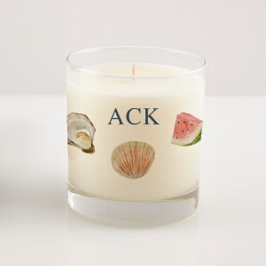 May include: A clear glass candle with white wax featuring the text ACK in dark blue letters, accompanied by watercolor-style illustrations of an oyster shell, a scallop shell, and a slice of watermelon printed on the front of the glass.